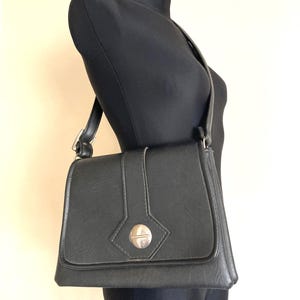 Pode incluir: Uma bolsa de ombro preta com fecho de aba e fecho prateado. A bolsa &eacute; exibida em um manequim preto. A al&ccedil;a &eacute; ajust&aacute;vel e a bolsa parece ser feita de um material semelhante ao couro. A bolsa tem formato retangular.