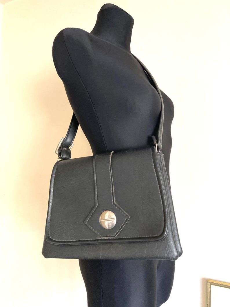 Pode incluir: Uma bolsa de ombro preta com fecho de aba e fecho prateado. A bolsa &eacute; exibida em um manequim preto. A al&ccedil;a &eacute; ajust&aacute;vel e a bolsa parece ser feita de um material semelhante ao couro. A bolsa tem formato retangular.