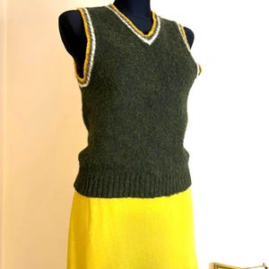 May include: A dark green, V-neck sweater vest with white and yellow trim, displayed on a black mannequin. The vest is paired with a bright yellow skirt. The outfit is a vintage style.