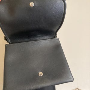 Pode incluir: Bolsa de m&atilde;o preta em couro sint&eacute;tico com fecho de aba e ferragens prateadas. A bolsa est&aacute; aberta, revelando o interior. A bolsa tem formato retangular com superf&iacute;cie texturizada. A bolsa &eacute; mostrada em um fundo neutro.