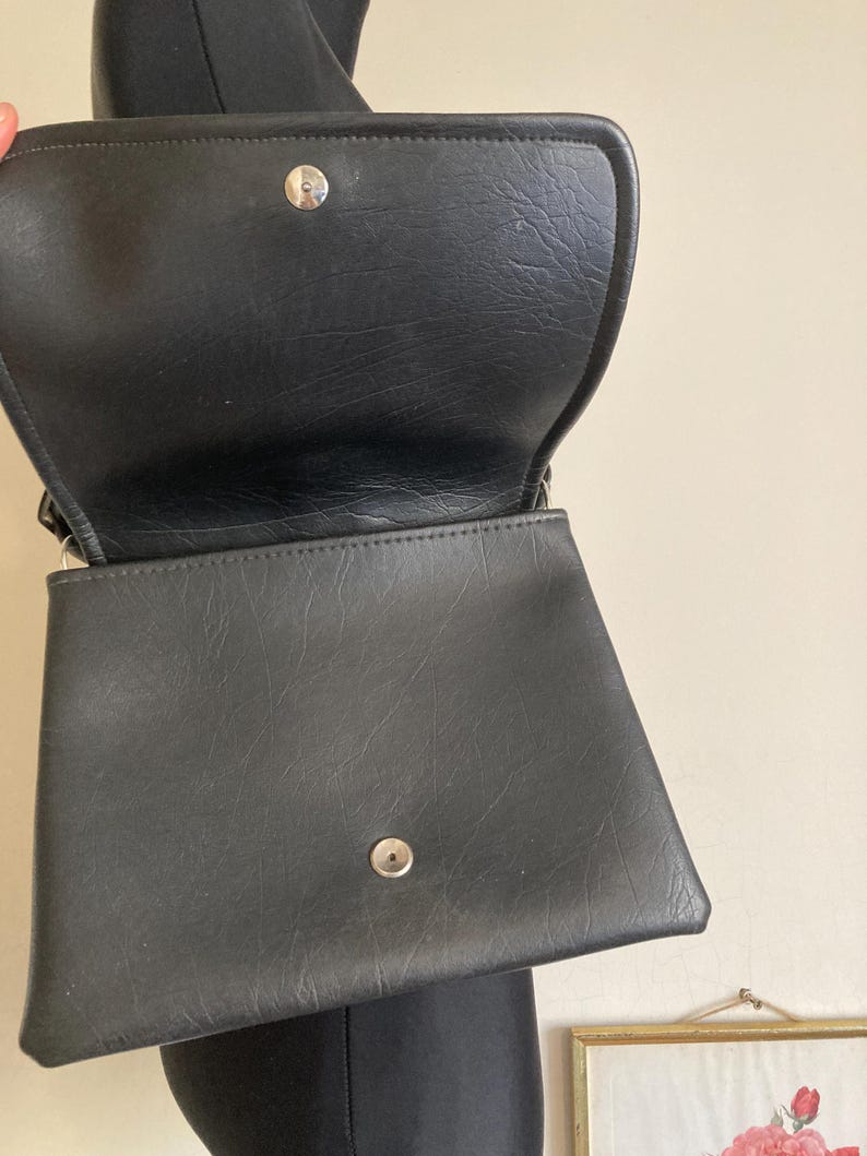 Pode incluir: Bolsa de m&atilde;o preta em couro sint&eacute;tico com fecho de aba e ferragens prateadas. A bolsa est&aacute; aberta, revelando o interior. A bolsa tem formato retangular com superf&iacute;cie texturizada. A bolsa &eacute; mostrada em um fundo neutro.