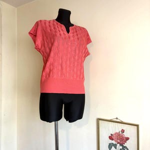 May include: A coral-coloured short-sleeved sweater with a textured pattern and a v-neck is displayed on a black mannequin. The sweater has a ribbed hem and is paired with black shorts. A framed botanical print hangs on the wall.