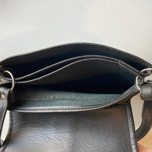 Pode incluir: Bolsa transversal de couro preta com a parte superior aberta, revelando v&aacute;rios compartimentos internos. A bolsa apresenta an&eacute;is de metal prateado e uma al&ccedil;a para transporte. O couro tem uma apar&ecirc;ncia texturizada.