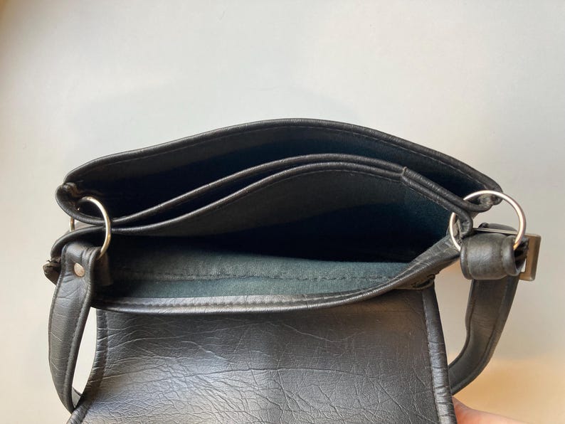 Pode incluir: Bolsa transversal de couro preta com a parte superior aberta, revelando v&aacute;rios compartimentos internos. A bolsa apresenta an&eacute;is de metal prateado e uma al&ccedil;a para transporte. O couro tem uma apar&ecirc;ncia texturizada.