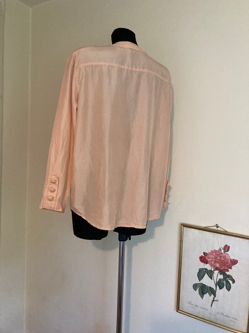 May include: A pale pink long-sleeved button-up shirt with a mandarin collar. The shirt is made of a silky material and has three buttons on each cuff.