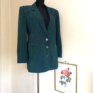 Puede incluir: Una chaqueta de color verde oscuro con cierre de un solo botón y dos bolsillos con solapa. La chaqueta está en un maniquí blanco.