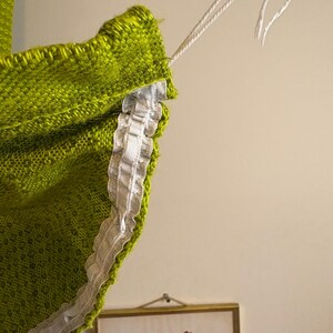 May include: Close-up of a lime green knitted textile with a white lace trim. White string is attached to the top edge. The textile has a textured appearance. A framed floral print is visible in the background.