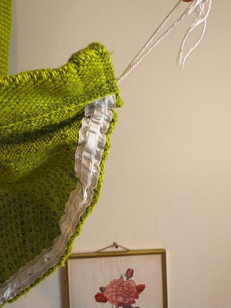 May include: Close-up of a lime green knitted textile with a white lace trim. White string is attached to the top edge. The textile has a textured appearance. A framed floral print is visible in the background.