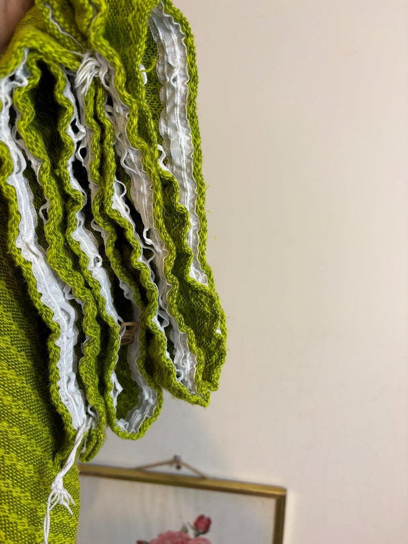 May include: Close-up of a lime green knitted fabric with white ruffled trim. The fabric has a textured appearance and is likely part of a garment. The white trim adds a decorative element to the edge of the fabric.