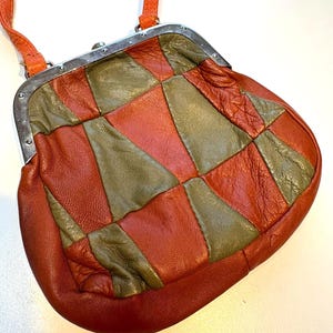 Peut inclure: Un sac à main vintage en cuir patchwork avec un fermoir argenté et une bandoulière orange. Le sac présente un motif géométrique de pièces de cuir rouge-orange et vert olive. Le sac a une forme arrondie et une conception structurée.