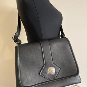 Pode incluir: Uma bolsa de ombro preta com fecho de aba e um fecho circular prateado. A bolsa tem formato retangular e uma al&ccedil;a longa. A bolsa &eacute; exibida em um manequim preto.