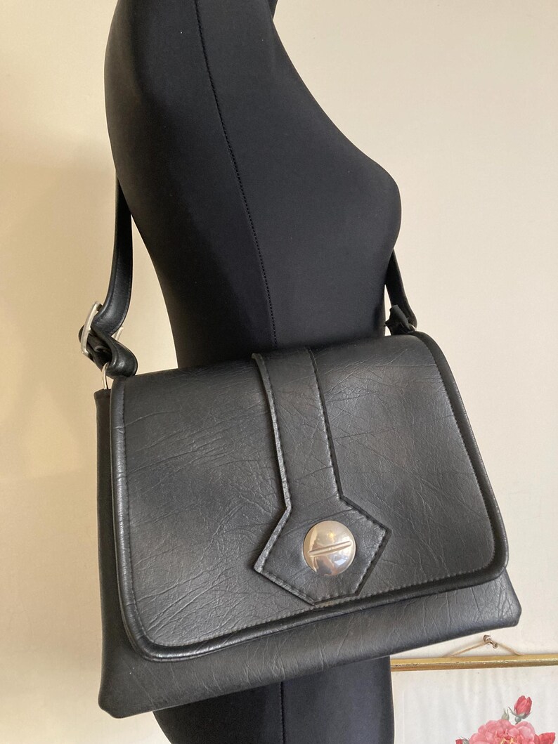 Pode incluir: Uma bolsa de ombro preta com fecho de aba e um fecho circular prateado. A bolsa tem formato retangular e uma al&ccedil;a longa. A bolsa &eacute; exibida em um manequim preto.