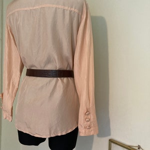 May include: A pale pink, long-sleeved blouse with a button-up front and a brown leather belt.