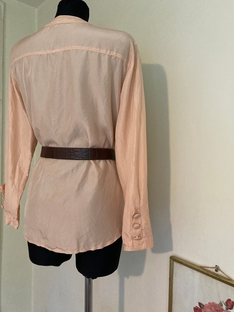 May include: A pale pink, long-sleeved blouse with a button-up front and a brown leather belt.