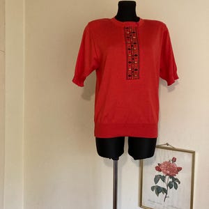 May include: A red short-sleeved sweater with a geometric pattern down the front. The sweater is on a mannequin.
