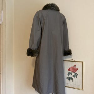 May include: A gray long coat with a faux fur collar and cuffs. The coat is hanging on a mannequin against a white wall.