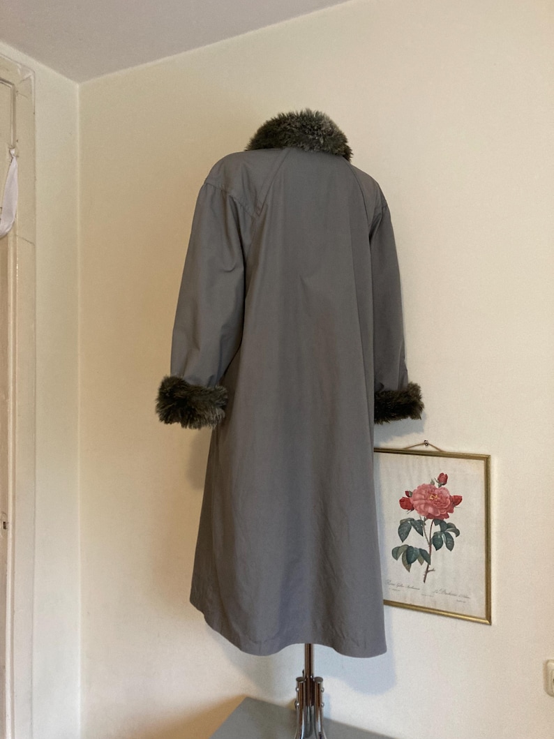 May include: A gray long coat with a faux fur collar and cuffs. The coat is hanging on a mannequin against a white wall.