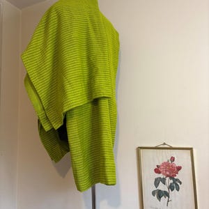 May include: A bright green, textured shawl draped over a metal clothing rack. The shawl has a woven pattern. A framed botanical print of a pink rose hangs on the white wall in the background.