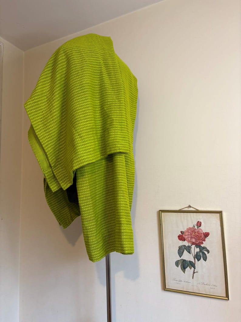 May include: A bright green, textured shawl draped over a metal clothing rack. The shawl has a woven pattern. A framed botanical print of a pink rose hangs on the white wall in the background.
