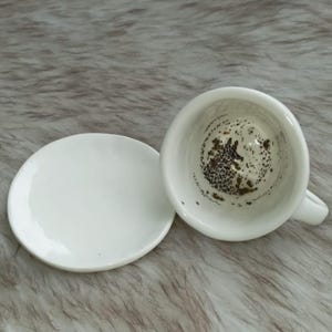 May include: A white ceramic teacup and saucer. The teacup is inverted, showing coffee grounds inside. The saucer is round and plain. The set rests on a textured, light-coloured surface.
