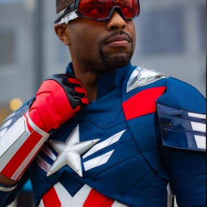 Pode incluir: Um traje inspirado no Capit&atilde;o Am&eacute;rica, com um esquema de cores azul e vermelho e uma estrela branca no peito. A pessoa tamb&eacute;m usa &oacute;culos vermelhos e luvas vermelhas e brancas. O traje tem detalhes prateados.