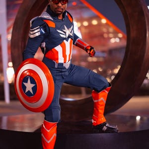 Pode incluir: Uma pessoa com uma fantasia inspirada no Capit&atilde;o Am&eacute;rica, segurando um escudo com uma estrela. A roupa &eacute; principalmente azul marinho, vermelha e branca, com um emblema de estrela no peito. A pessoa est&aacute; usando &oacute;culos vermelhos e botas laranja.