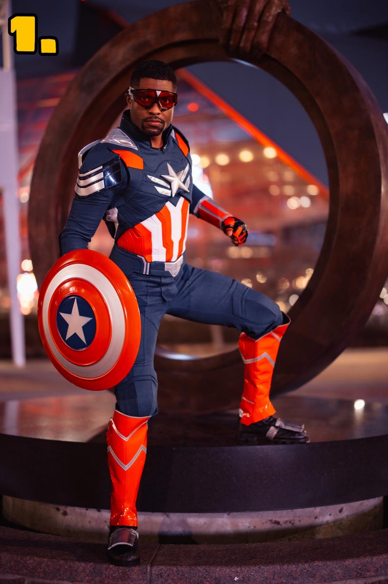 Pode incluir: Uma pessoa com uma fantasia inspirada no Capit&atilde;o Am&eacute;rica, segurando um escudo com uma estrela. A roupa &eacute; principalmente azul marinho, vermelha e branca, com um emblema de estrela no peito. A pessoa est&aacute; usando &oacute;culos vermelhos e botas laranja.