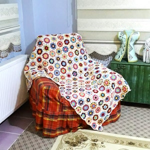 May include: A crocheted throw blanket with a floral pattern in shades of white, pink, blue, and brown. The blanket is draped over a red and black plaid armchair.