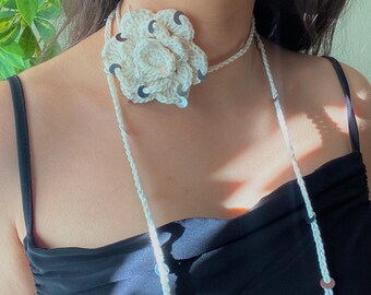 Può includere: Collana girocollo a fiori all'uncinetto color crema con paillettes argentate. Il fiore è il punto focale, con un delicato cordino intrecciato che si allaccia sul retro. La collana è indossata sul collo di una persona.