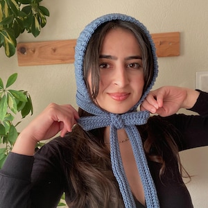 May include: A light blue knitted bonnet with long ties, knotted in a bow under the chin. The bonnet is a solid colour and appears to be handmade. The bonnet covers the head and frames the face, with the ties hanging down.