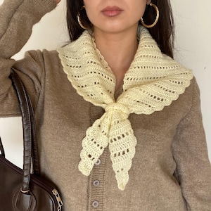 May include: A pale yellow triangular crochet scarf featuring an openwork stitch pattern with scalloped edges, tied in a knot at the front. The scarf is worn over a brown long-sleeved button-down cardigan, paired with a dark brown shoulder bag.