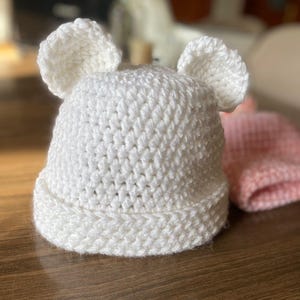 Pode incluir: Um chapéu branco de crochê feito à mão com duas orelhas pequenas e arredondadas no topo. O chapéu tem uma aba e é feito de um fio grosso e texturizado. Um chapéu rosa de crochê é parcialmente visível no fundo. Este chapéu é para bebês e crianças pequenas.