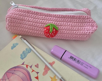 Puede incluir: Un estuche para lápices de crochet rosa con una cremallera blanca y una aplicación de fresa roja. El estuche está sobre una mesa con un lápiz blanco y un resaltador morado.