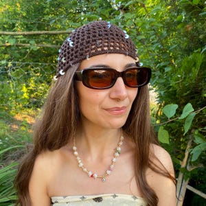 May include: A brown crocheted hat with silver sequins is worn with brown sunglasses and a beaded necklace. The necklace has white, red, and green beads, and a small blue eye charm. The background is a green, leafy outdoor setting.