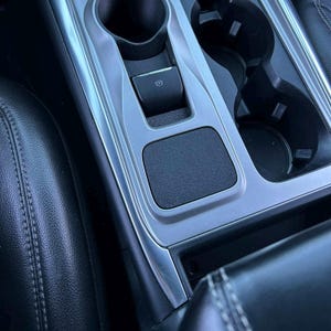 May include: Interior car console with a silver trim. Features include two cup holders, a parking brake button, and a small, textured pad. The console is set against a black leather seat.