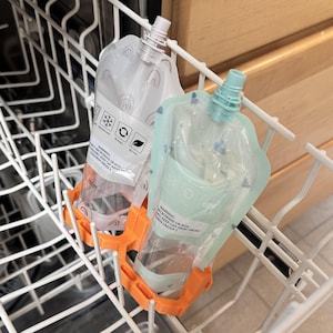 May include: Two clear, reusable food pouches with white tops and orange holders are in a dishwasher rack. One pouch has a rainbow design, the other is light green. Text on the pouches includes warnings and recycling symbols.