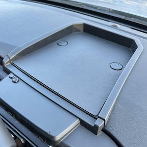 以下が含まれることがあります： 2つの楕円形のくぼみがある黒いプラスチック製のダッシュボードトレイ。トレイは長方形で、縁が盛り上がっており、テクスチャ加工されたダッシュボードに取り付けられています。ボタンが付いた、より小さな長方形の部分が左側に表示されます。