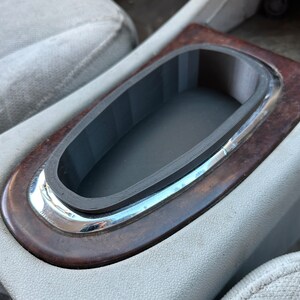 May include: A close-up of a car's center console with a black rubber cup holder insert. The cup holder is surrounded by a silver trim and a dark wood-grain finish.