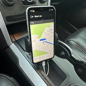 May include: A black phone holder mounted in a car's centre console. The phone is displaying a map app with the text "Start on" and a route with an estimated arrival time of 11:45. The phone is connected to a charging cable.