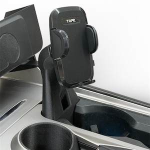 Könnte beinhalten: Schwarzer TOPK-Telefonhalter, montiert in der Konsole eines Autos. Der Halter hat verstellbare Arme und befindet sich über einem Getränkehalter. Die Konsole ist schwarz und silberfarben.
