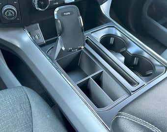 May include: A gray car interior with a phone holder mounted in the center console. The phone holder is black and has the word "TOPK" printed on it. The console has a cup holder and a storage compartment.
