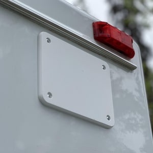 Puede incluir: Primer plano de una placa rectangular blanca con esquinas redondeadas, asegurada por cuatro tornillos, montada sobre una superficie gris claro. Una luz trasera roja es visible encima de la placa. Imagen de un vehículo.