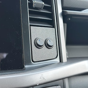 May include: A close-up of a car's interior, featuring a textured black panel with two round, black switches. Each switch has a blue indicator light. The panel is mounted near the air vent and dashboard.
