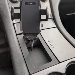 以下が含まれることがあります： 前面にTOPKのブランド名が入った黒い車の電話ホルダー。電話ホルダーは車のセンターコンソールに取り付けられています。コンソールは黒とグレーで、カップホルダーとエアコンのコントロールが付いています。