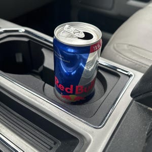 May include: A silver can of Red Bull energy drink in a car cup holder. The can is partially empty and has a blue and red label with the text "Red Bull" in white.