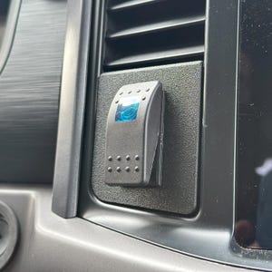 Può includere: Un interruttore a levetta nero con una luce LED blu. L'interruttore è montato su un pannello in plastica nera.