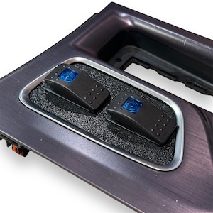 Könnte beinhalten: Zwei schwarze Kippschalter mit blauen LED-Leuchten, montiert auf einer gebürsteten Metallplatte. Die Schalter sind von einer silbernen Einfassung umgeben.