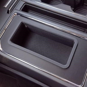 May include: A close-up of a car's center console, featuring a rectangular storage compartment with a textured, dark gray interior. The compartment is framed by a smooth, black surface and a silver trim.