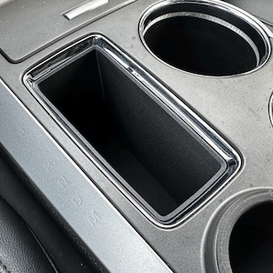May include: A close-up of a gray car interior with a rectangular storage compartment and two cup holders. The compartment is empty and has a silver trim. The cup holders are also empty and have a silver trim.
