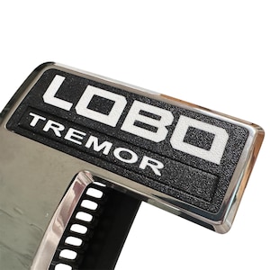 Può includere: Un'emblema cromato e nero con la parola "LOBO" in lettere bianche a blocchi sopra "TREMOR". L'emblema ha uno sfondo nero strutturato e un bordo cromato lucido. Il design è rettangolare.